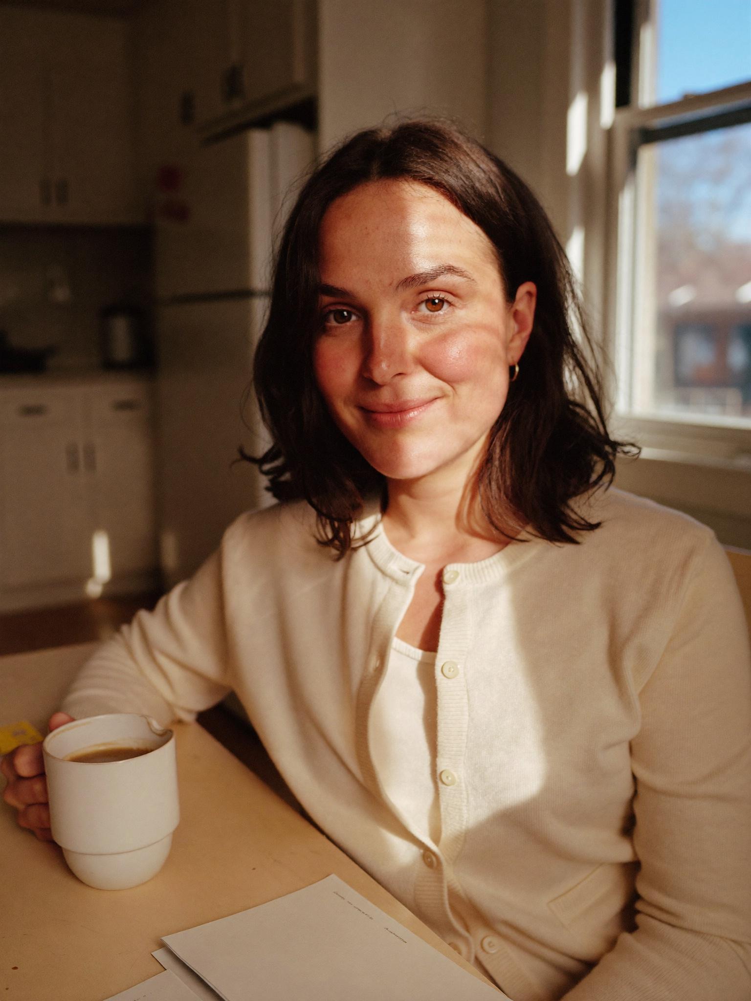 A young woman sitting at a sunlit kitchen table with a coffee mug.