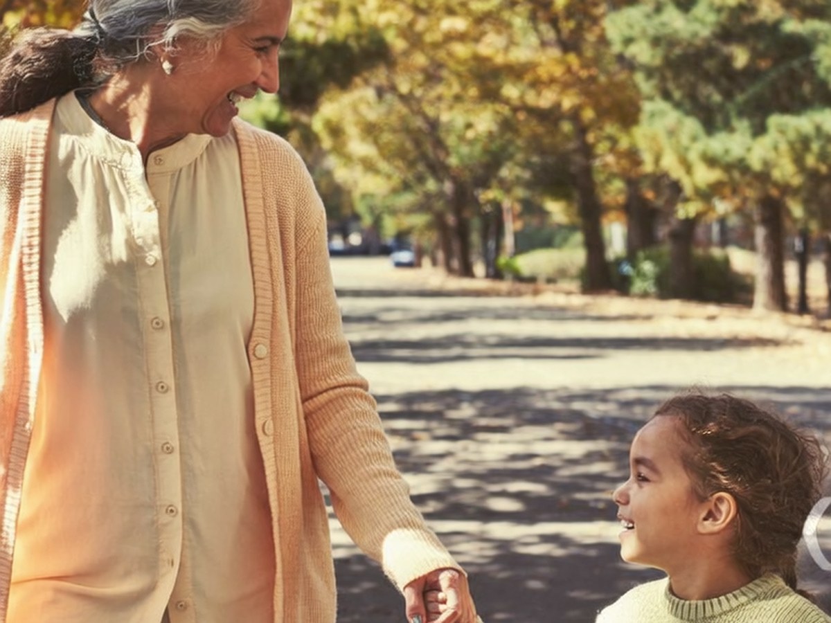 A grandmother walking on a tree-lined park path holding hands with her young granddaughter.