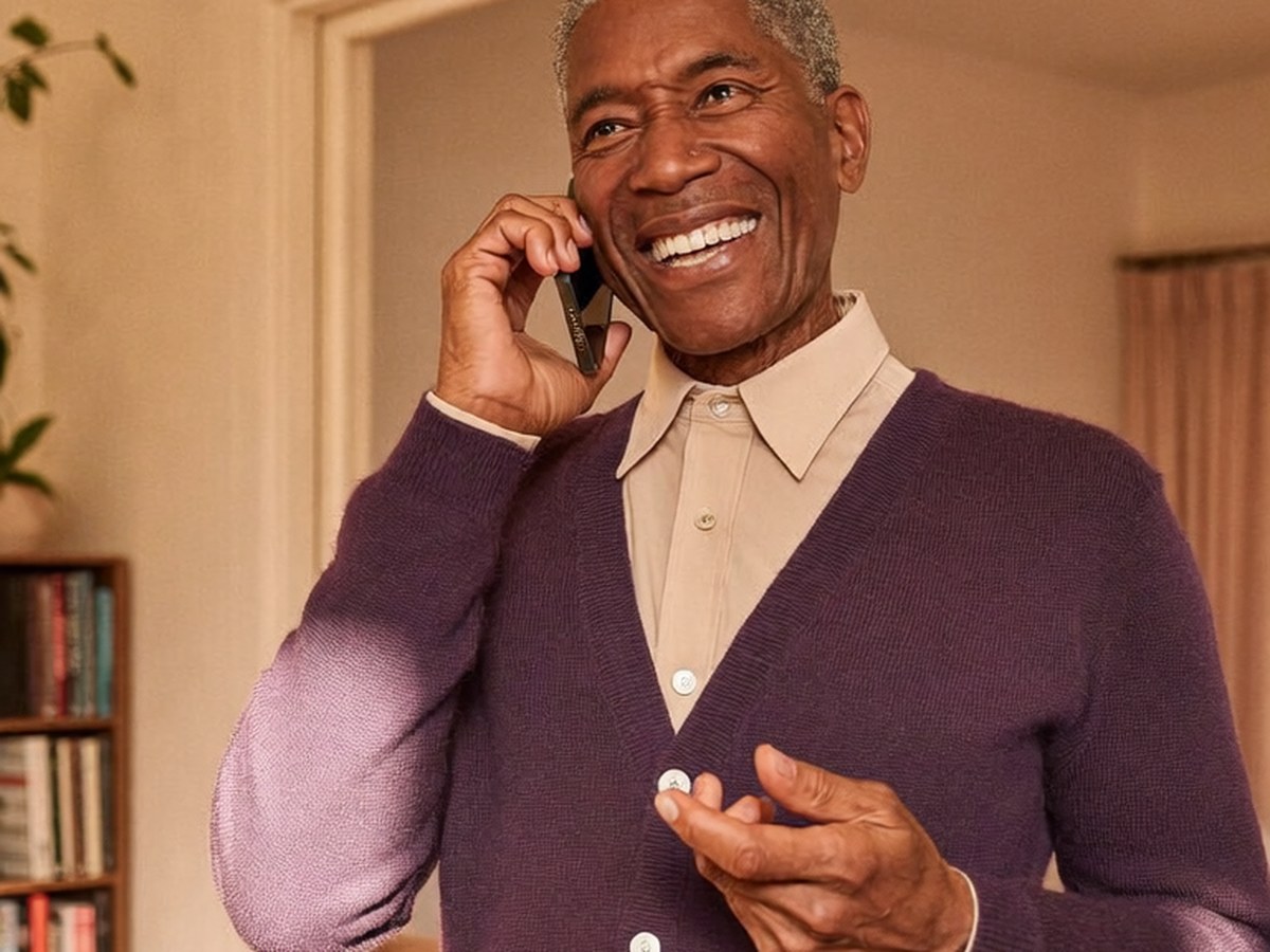 An older man in his living room laughing on a phone call.